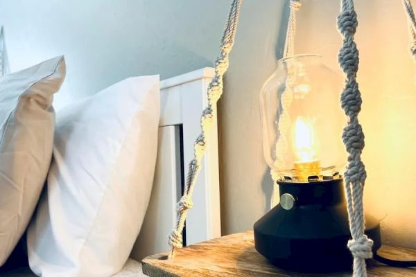 A cozy bedroom scene with pillows on a bed, a lit lamp on a wooden side table, and decorative knotted ropes hanging down.