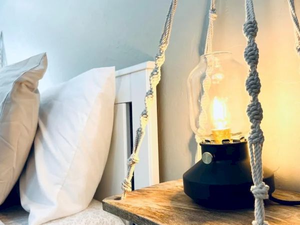 A cozy bedroom scene with pillows on a bed, a lit lamp on a wooden side table, and decorative knotted ropes hanging down.