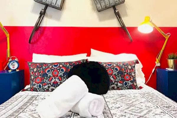 A bed with decorative pillows and rolled white towels is flanked by yellow lamps; flower pots and bicycle elements hang on the wall above the bed.