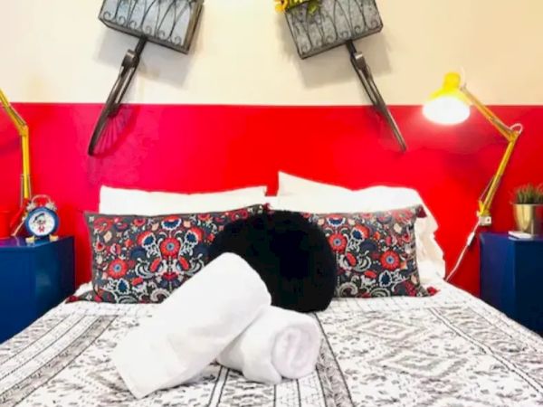 A bed with decorative pillows and rolled white towels is flanked by yellow lamps; flower pots and bicycle elements hang on the wall above the bed.