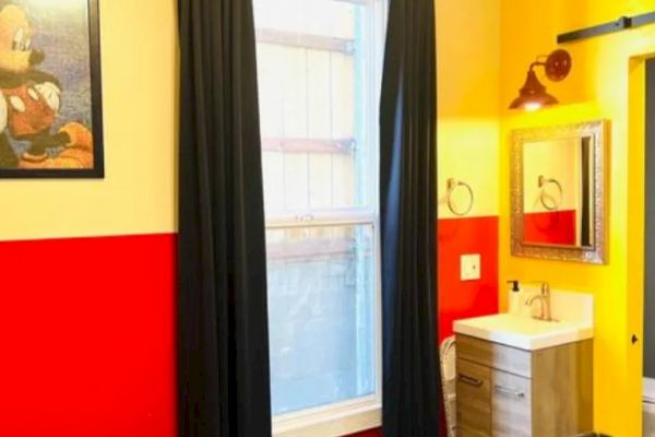 A room with red and yellow walls, black curtains, a window, a framed picture, a sink with a mirror, and a trash bin.