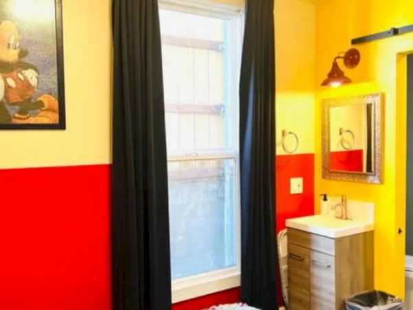 A room with red and yellow walls, black curtains, a window, a framed picture, a sink with a mirror, and a trash bin.