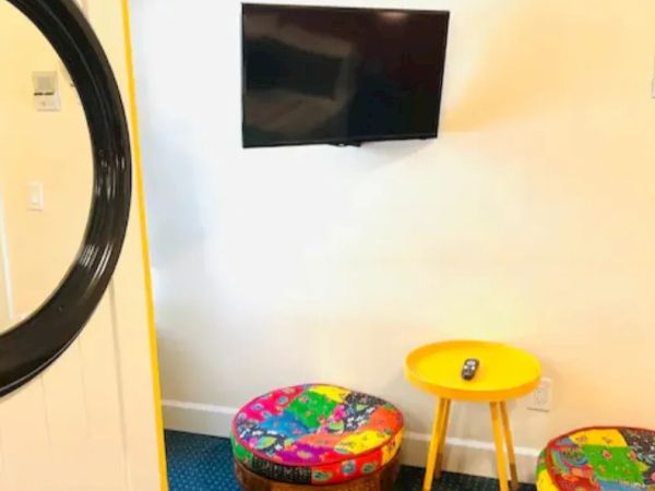 The image features a small seating area with two colorful ottomans, a yellow side table, a mounted TV, and part of a mirror on the left.