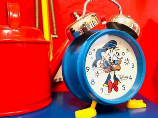 A blue alarm clock with a cartoon character, likely from Disney, next to a red container on a blue surface.