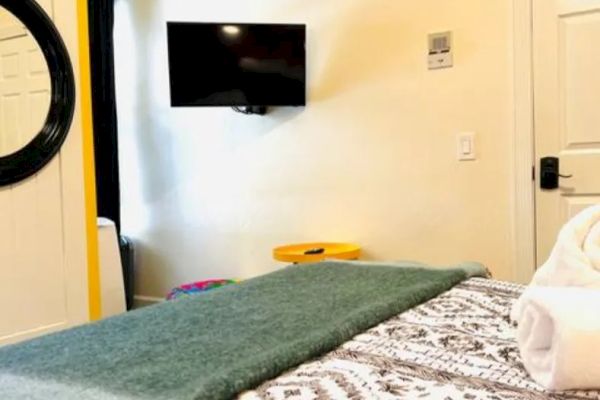 A cozy bedroom with a patterned bedspread, a green throw, a wall-mounted TV, a round mirror, and neatly rolled towels on the bed.