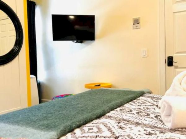 A cozy bedroom with a patterned bedspread, a green throw, a wall-mounted TV, a round mirror, and neatly rolled towels on the bed.
