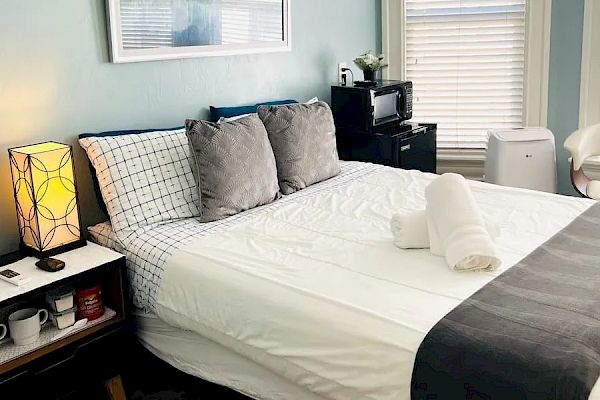 A neatly made bed with gray pillows and a dark throw, a bedside table with a lamp, and a small kitchen area against a light blue wall.