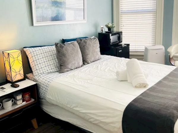 A neatly made bed with gray pillows and a dark throw, a bedside table with a lamp, and a small kitchen area against a light blue wall.