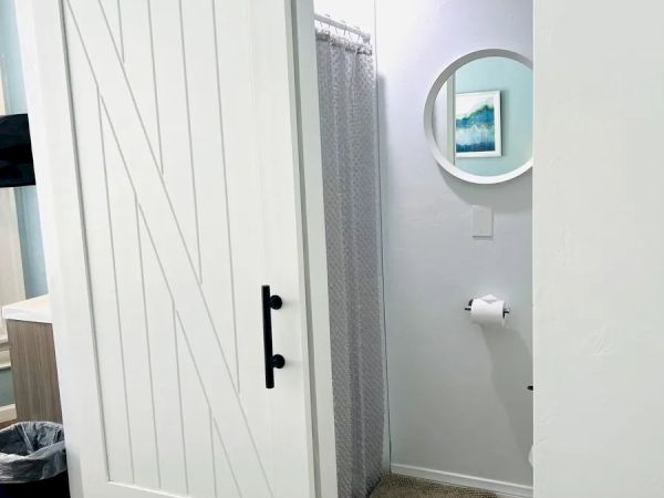 The image shows a sliding barn door leading to a small bathroom with a round mirror, a framed picture, a shower curtain, a rug, and a toilet paper holder.