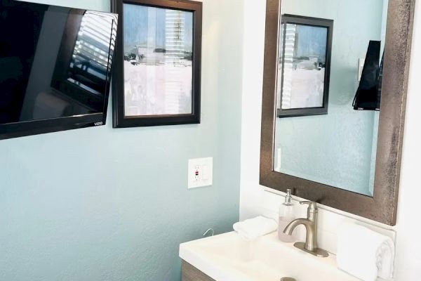 A bathroom corner with a wall-mounted TV, two framed pictures, a large mirror above a sink on a wooden cabinet, with a soap dispenser and towel.