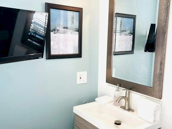 A bathroom corner with a wall-mounted TV, two framed pictures, a large mirror above a sink on a wooden cabinet, with a soap dispenser and towel.