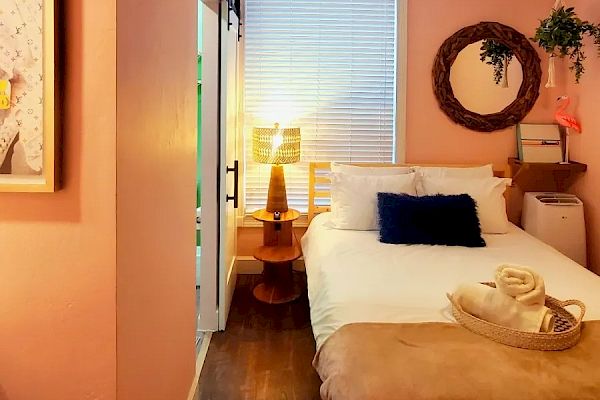 The image shows a cozy bedroom with pink walls, a bed, a bedside table, a lamp, a circular mirror, plants, and a ceiling fan.