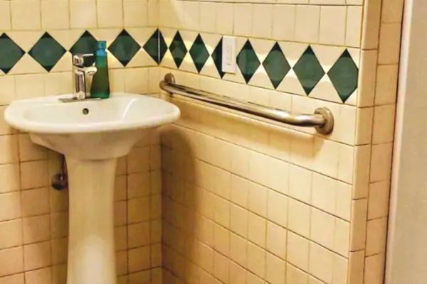 A bathroom with a pedestal sink, green diamond-patterned tiles, a mirror, a liquid soap dispenser, and a metal grab bar on the wall.