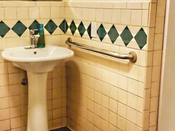 A bathroom with a pedestal sink, green diamond-patterned tiles, a mirror, a liquid soap dispenser, and a metal grab bar on the wall.