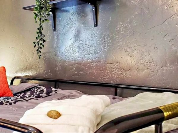 A cozy bedroom corner featuring a bed, two white towels, a red pillow, and a shelf with potted plants on a textured wall.