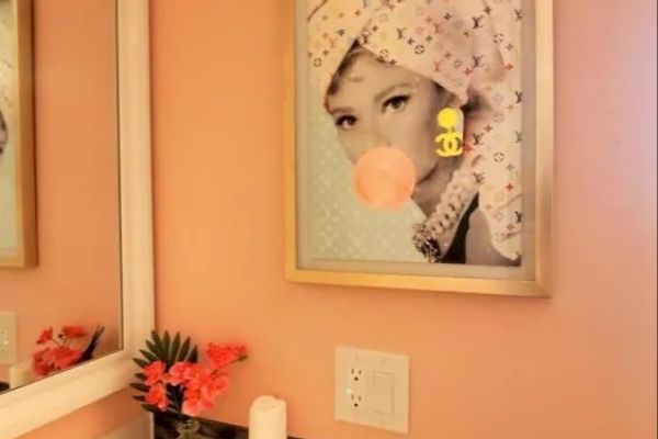 The image shows a bathroom with light pink walls, featuring a framed picture of a woman with a towel and bubblegum. There's a mirror and a counter with flowers.