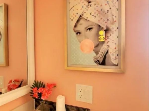 The image shows a bathroom with light pink walls, featuring a framed picture of a woman with a towel and bubblegum. There's a mirror and a counter with flowers.