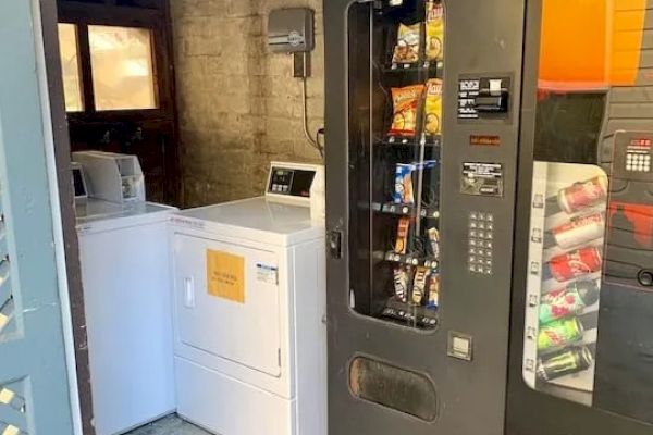 The image shows a small room with a washing machine, a dryer, a snack vending machine, and a drink vending machine.