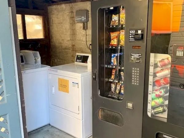 The image shows a small room with a washing machine, a dryer, a snack vending machine, and a drink vending machine.
