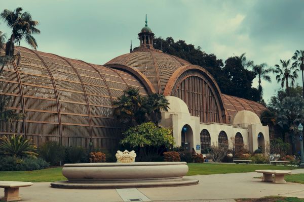 A large, historic greenhouse building with a domed roof and arched windows, surrounded by greenery and palm trees, featuring a central fountain.