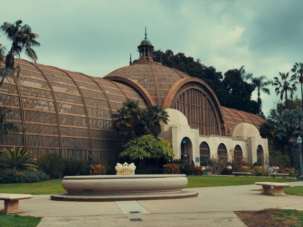 A large, historic greenhouse building with a domed roof and arched windows, surrounded by greenery and palm trees, featuring a central fountain.