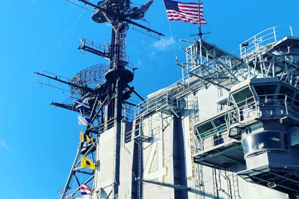 The image depicts a naval ship superstructure, featuring an antenna mast with an American flag and signal flags, set against a clear blue sky.