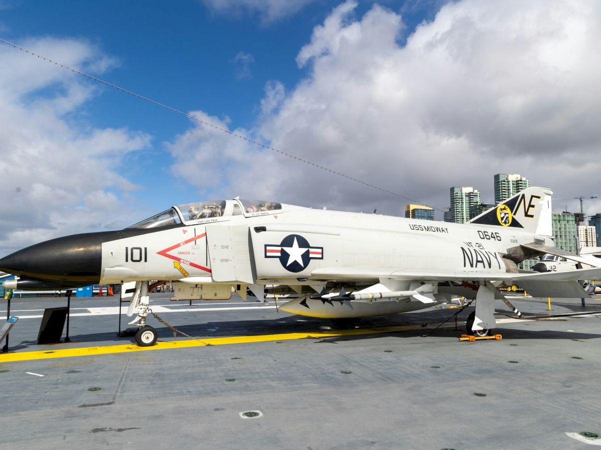 The image shows a U.S. Navy jet aircraft, marked with the insignia of the Navy, parked on the deck of a naval vessel under a partly cloudy sky.