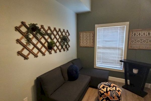 A cozy room with a dark sofa, patterned ottoman, wall decor, and a window with blinds. It has wooden accents and a plant on the wall.