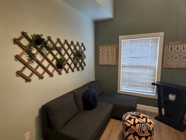 A cozy room with a dark sofa, patterned ottoman, wall decor, and a window with blinds. It has wooden accents and a plant on the wall.