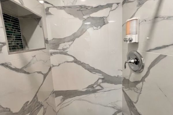 A modern shower with marble-patterned tiles, a built-in shelf, and a wall-mounted soap dispenser.