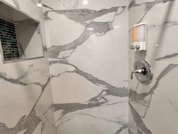 A modern shower with marble-patterned tiles, a built-in shelf, and a wall-mounted soap dispenser.