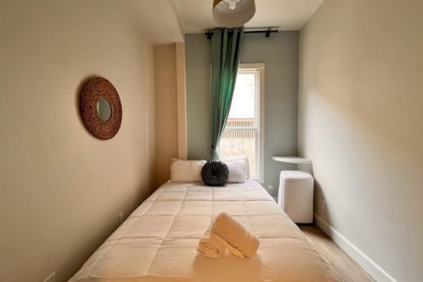 A small, cozy bedroom with a neatly made bed, rolled towels, a mirror on the wall, a small window with green curtains, and minimalistic furniture.