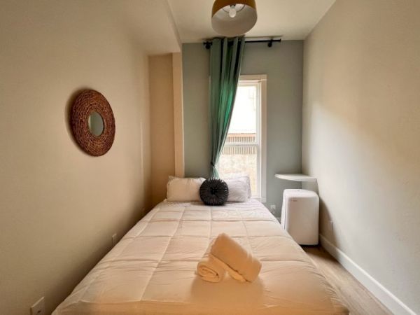 A small, cozy bedroom with a neatly made bed, rolled towels, a mirror on the wall, a small window with green curtains, and minimalistic furniture.