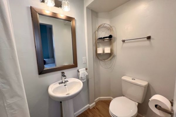 A small bathroom with a pedestal sink, framed mirror, toilet, towel and toilet paper holders, and a wall-mounted shelf holding toiletries.