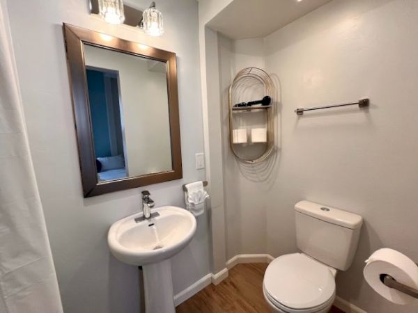 A small bathroom with a pedestal sink, framed mirror, toilet, towel and toilet paper holders, and a wall-mounted shelf holding toiletries.