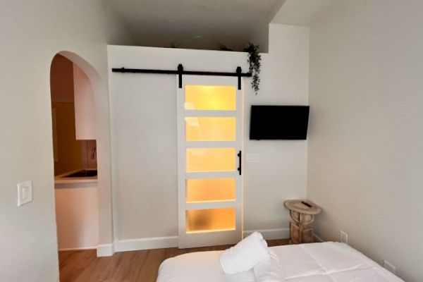 The image shows a cozy bedroom with a sliding barn door, a wall-mounted TV, and a small table. An archway leads to a kitchenette area.