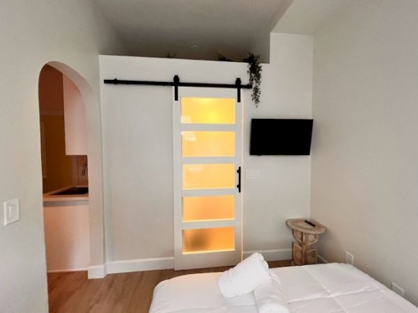 The image shows a cozy bedroom with a sliding barn door, a wall-mounted TV, and a small table. An archway leads to a kitchenette area.