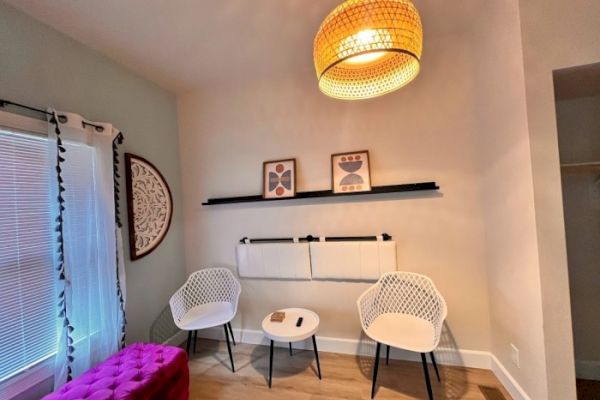 A cozy room with two white chairs, a small table, a purple footrest, wall art, a woven pendant light, and window blinds with drapes.