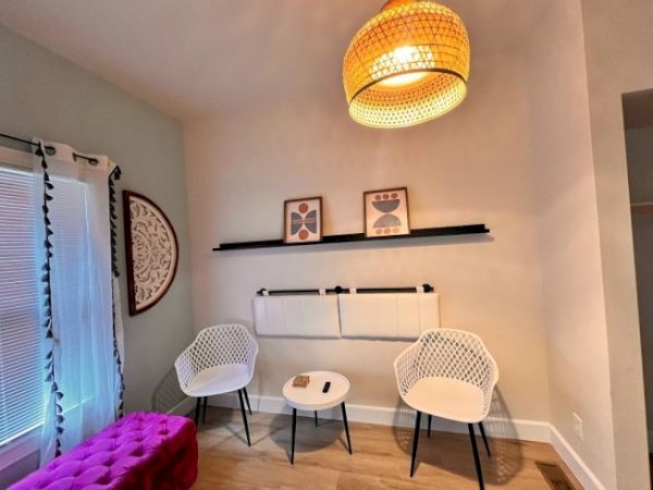 A cozy room with two white chairs, a small table, a purple footrest, wall art, a woven pendant light, and window blinds with drapes.