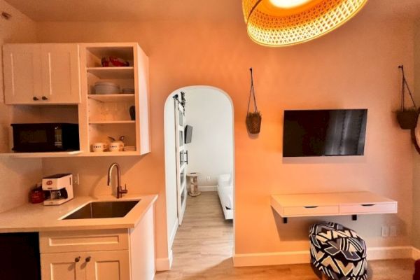 A small kitchen area with a sink, microwave, and open shelving, next to an open doorway leading to a bedroom; TV mounted on the wall.