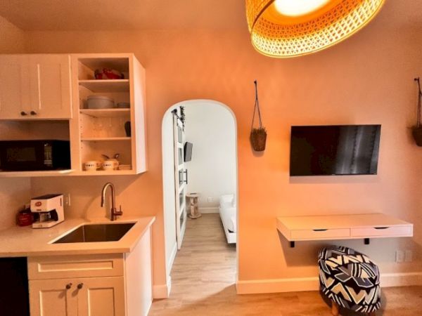 A small kitchen area with a sink, microwave, and open shelving, next to an open doorway leading to a bedroom; TV mounted on the wall.