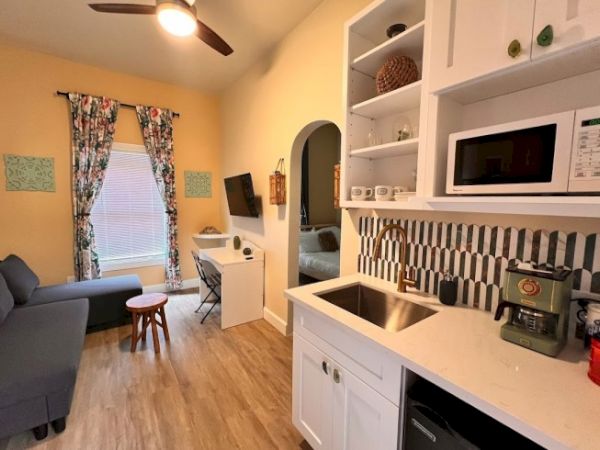 A cozy living space with a sectional sofa, a wall-mounted TV, a computer desk, and a kitchenette with a microwave, sink, and coffee maker.