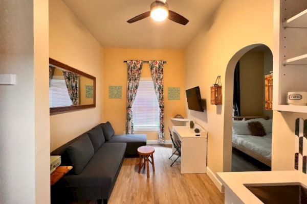 A cozy living area with a sofa, coffee table, wall-mounted TV, desk, ceiling fan, and entrance to a bedroom, in a well-lit room with wooden floors.