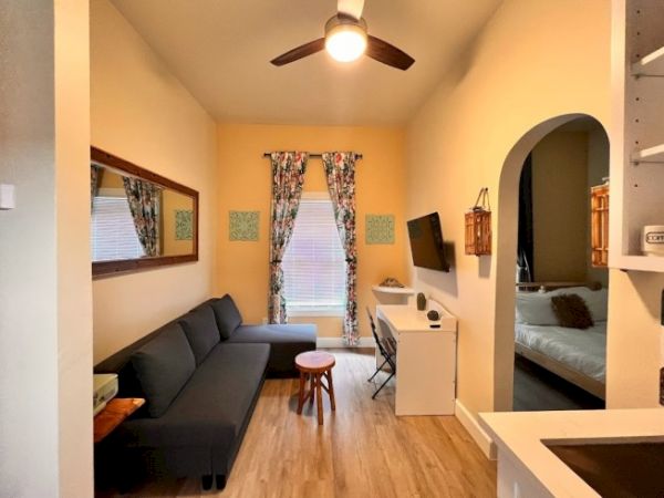 A cozy living area with a sofa, coffee table, wall-mounted TV, desk, ceiling fan, and entrance to a bedroom, in a well-lit room with wooden floors.