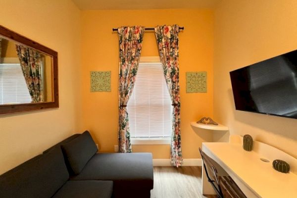 A cozy room with a grey sofa, wall-mounted TV, desk with cacti decor, window with floral curtains, wall mirror, and yellow walls.