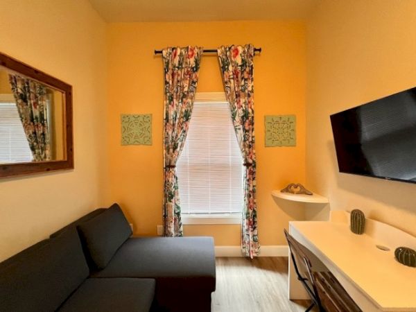 A cozy room with a grey sofa, wall-mounted TV, desk with cacti decor, window with floral curtains, wall mirror, and yellow walls.
