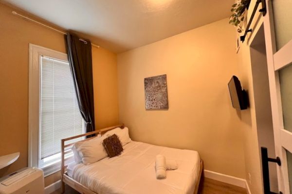 The image shows a cozy bedroom with a single bed, wall-mounted TV, window with dark curtains, white bedding, and a small piece of artwork on the wall.