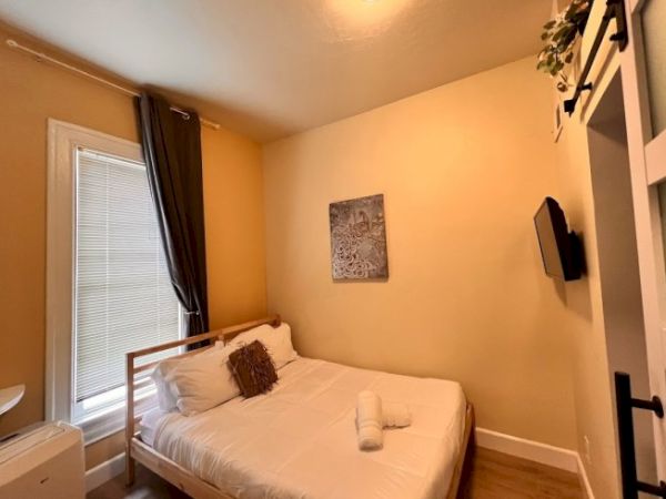 The image shows a cozy bedroom with a single bed, wall-mounted TV, window with dark curtains, white bedding, and a small piece of artwork on the wall.