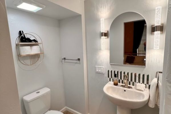 A modern bathroom with a wall-mounted sink, mirror, light fixtures, toilet, and a circular shelf unit holding toiletries and towels above the toilet.