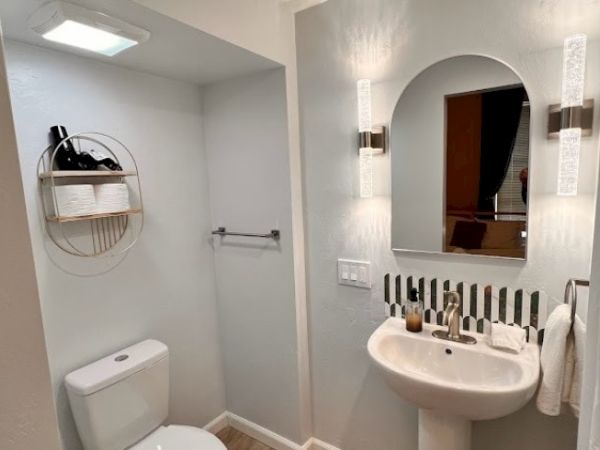A modern bathroom with a wall-mounted sink, mirror, light fixtures, toilet, and a circular shelf unit holding toiletries and towels above the toilet.